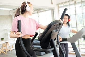 Cheerful friends are running together on a running machine at a sports center by young lady smile and looking at senior lady. Both wear headphone during running on treadmill and look happy.