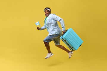 Happy African-American man in shorts and a cap jumps with a suitcase and tickets in his hands on...