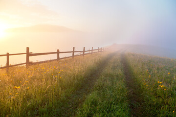 Sunrise over mountain meadow with wooden fence and old trail, a path to foggy background. Ukraine, carpathians.