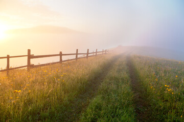 Fototapeta premium Sunrise over mountain meadow with wooden fence and old trail, a path to foggy background. Ukraine, carpathians.