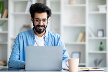Smiling young indian man working on laptop at home office