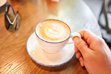 A cup of hot cappuccino on the table in the coworking space of the coffee shop. A cup of coffee on the veranda on a sunny day. Coffee break at the cafe. Close-up