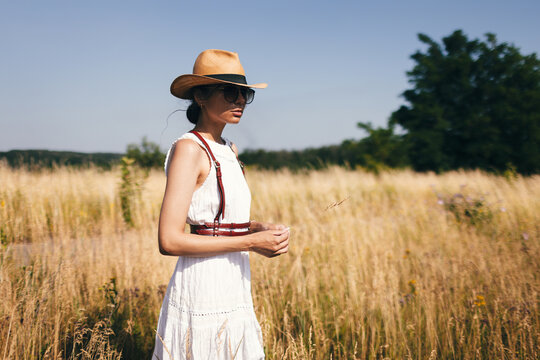 Spirit of freedom. An attractive boho girl in blouse, hat and sunglasses standing on the field on the background of a blue sky. Summer vacation, traveling. Bohemian, modern hippie style.