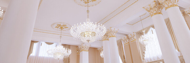 Beautiful vintage luxury white ceiling with crystal chandeliers and golden stucco. White columns of the wedding palace decorated with stucco.Banner for website header design with copy space.