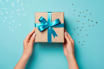  First person top view photo of hands unpacking craft paper gift box with blue satin ribbon bow over shiny sequins on isolated pastel blue background