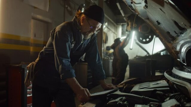 Medium long shot of mature Caucasian specialist working in auto service garage using digital tablet to identify problem with engineMedium long shot of mature Caucasian specialist working in auto servi