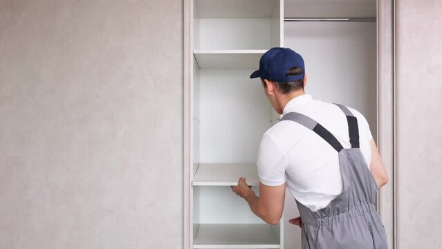 Male employee in work uniform assembles light wardrobe attaching shelves. Furniture assembly service concept. Man of all trades assembles closet