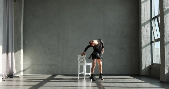 Active Caucasian dancer unleashes his energy and grooves with passion near a chair in high heels at a vibrant dance studio. His powerful moves, combined with the elegance of high heels.