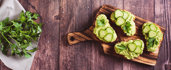 Crostini with avocado puree, cucumbers and herbs on a wooden board top view web banner