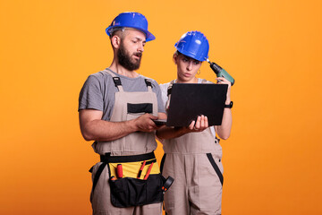 Professional builders in studio holding laptop, looking at building blueprint for new renovation project. Renovators wearing renovating uniform making decisions together as team.