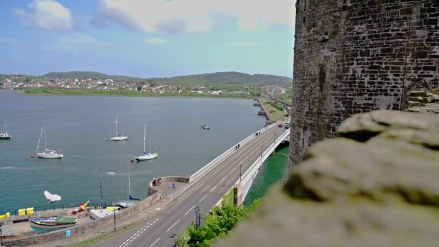 High aerial view of River Conwy, boats, cars on bridge and a castle tower, from the ledge of a stone wall at Conwy Castle, on a spring afternoon, in Conwy, Wales, UK