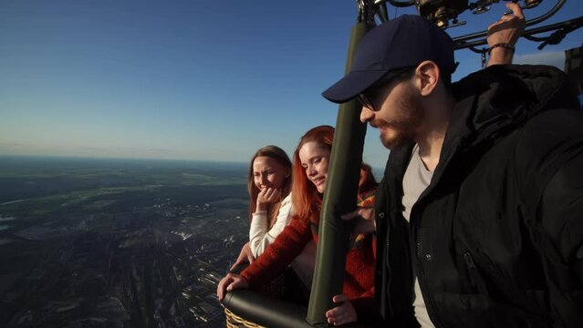 Man and two women are flying on hot air balloon and enjoying amazing view on green valleys. Male caucasian person is taking selfie of himself and his two female friends being inside basket of vehicle.