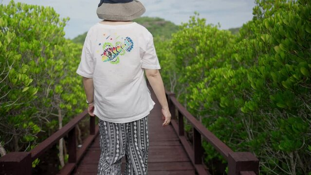 With a blue hat and a white t-shirt, an old Asian woman walks in an Asian forest, her hand reaching for the sky. The camera follows her capturing a captivating moment of joy and oneness with nature.