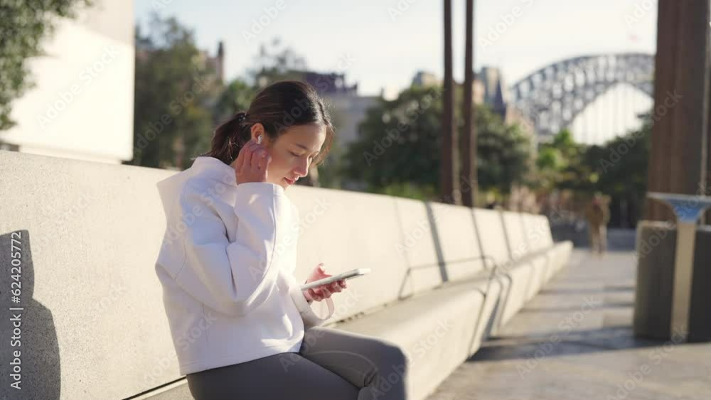 Wall mural Asian woman in sportswear listening to music from earphones with mobile phone app during jogging exercise at city street in the morning. Healthy girl do sport training running workout in the city.