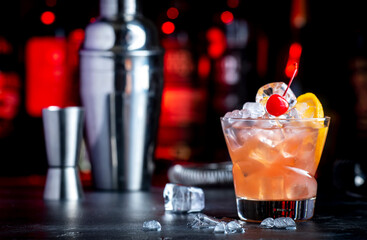 Matador alcoholic cocktail drink with scotch whiskey, white vermouth, liqueur, bitter, grenadine, orange juice, cocktail cherry and ice. Black bar counter background, steel bar tools and bottles