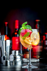 Rose sangria summer alcoholic drink with pink wine, peach, orange, lime and ice. Black bar counter background, steel bar tools and bottles