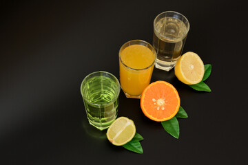 Three tall glasses with different citrus juices on a black background, next to the halves of ripe orange, lemon and lime with leaves.