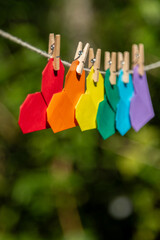 Varal com diversos corações de origami pendurados bem juntinhos com as cores da bandeira do orgulho gay em fundo de vegetação desfocado.