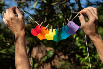 Mãos de mulher morena em meio à natureza pendurando um pequeno varal de corações de origami...