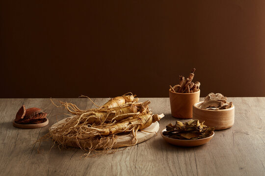 On the wooden table and brown background, chinese traditional medicine with fresh ginseng roots and some medicine herbs places on wooden plate and tray. Front view, herbs originating from the east