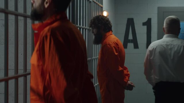 Two prisoners in orange uniforms stand facing the metal bars in front of prison cells. Prison officer with police baton walks, watches criminals in jail. Detention center or correctional facility.