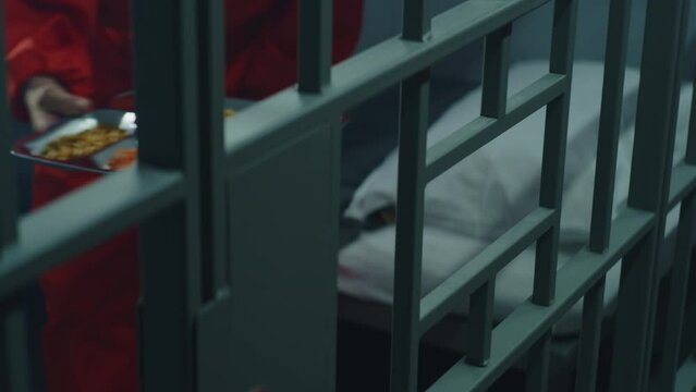 Close up of prison warden giving tray of food through metal bars to inmate. Elderly prisoner in orange uniform sits in prison cell, eats dinner. Serving imprisonment term in jail or detention center.