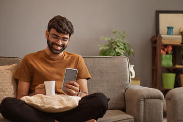 Young man drinking coffee and using mobile phone while sitting on sofa in living room
