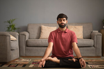 Young man meditating in living room - obrazy, fototapety, plakaty