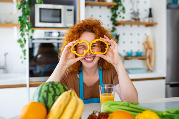 Photo of young woman smiling and holding pepper circles on her eyes while cooking salad with fresh...