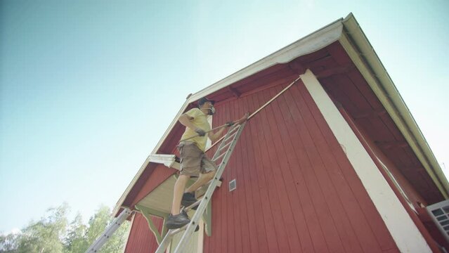 Man climbs ladder with long brush and removes peeling red paint from wooden home