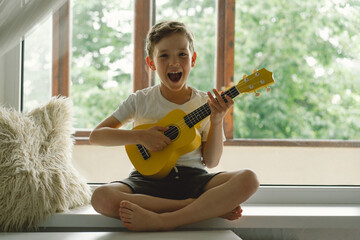 Cute boy learns to play the yellow Ukulele guitar on the windowsill near the window. Cozy home....
