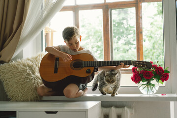 Cute boy learns to play the classical guitar on the windowsill near the window with cat. Cozy home....