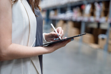 Young beautiful Asian manager stocking products in cardboard boxes on shelves in warehouse using digital tablet and pen.