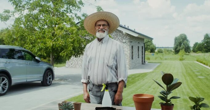 An elderly man with a beard, glasses and a hat, smiling looking at the camera, standing in the backyard of his house