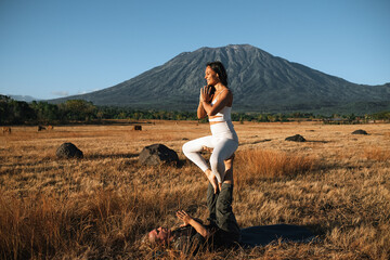 Naklejka na ściany i meble A couple of young people, a man and a woman, of European appearance, are engaged in acroyoga in nature, against the backdrop of Agung volcano, Bali.