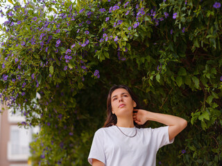 Beautiful young woman with dark curly hair and perfect skin wearing white tshirt posing near blooming flowers in garden. Nude make up. Allergy season, antihistamines. Aroma oils natural perfumes