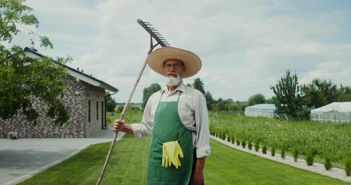 An elderly man with a gray beard, dressed in an apron and hat, smiles while looking at the camera standing in the backyard with a rake in his hand
