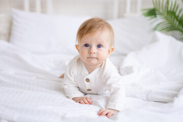 smiling baby 6 months old blond boy crawls on a white bed in a bright bedroom after sleeping in the...