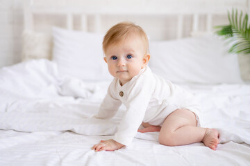 smiling baby 6 months old blond boy crawls on a white bed in a bright bedroom after sleeping in the morning in a cotton bodysuit, the concept of children's goods