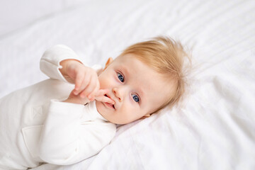 smiling baby 6 months old blond boy lies on a white bed in a bright bedroom and licks his finger in a cotton bodysuit, the concept of children's goods