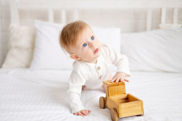 smiling baby 6 months old blond boy is sitting on a large bed in a bright bedroom and playing with a wooden toy car in a cotton bodysuit, the concept of children's goods