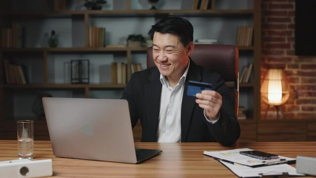 Cheerful asian employee having access to unlimited credit card and doing online shopping in comfy office. Smiling man dressed in black suit making payment on laptop via internet banking.
