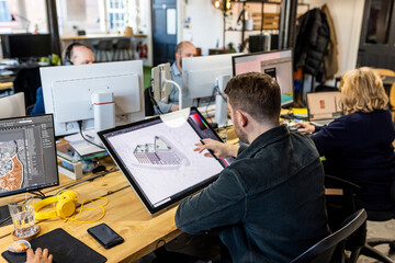 Architect working on graphics tablet with colleagues at desk in office