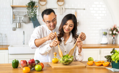 Young asian family couple having fun cooking and preparing cook vegan food healthy eat with fresh vegetable salad on counter in kitchen at home.Happy couple looking to preparing food