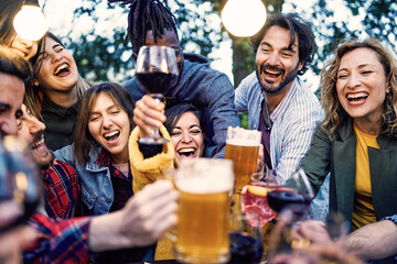 Multiracial Friends Celebrating Outdoors - Joyous group toasting with beer, wine, and sangria at an outdoor party at dusk, under hanging garden lights.