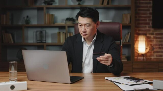 Company owner making online payment with credit card through wireless laptop in creative workplace. Asian boss with serious face expression purchasing goods on websites. Concept of internet banking. - Powered by Adobe