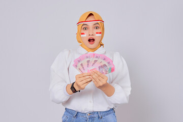 Shocked young Asian Muslim woman wearing hijab holding money Indonesian rupiah banknotes isolated over white background. Celebrate Indonesian independence day on 17 August