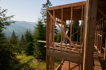 Fototapeta na wymiar Man and woman inspecting their future wooden frame house nestled in the mountains near forest. Youthful couple at construction site in early morning. Concept of contemporary ecological construction.