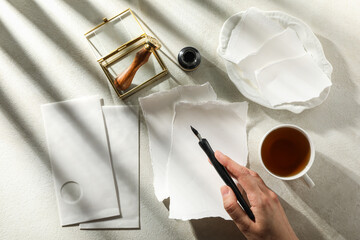 Sheets of paper, stamp, ink bottle and pen in hand, cup of tea on light background, top view