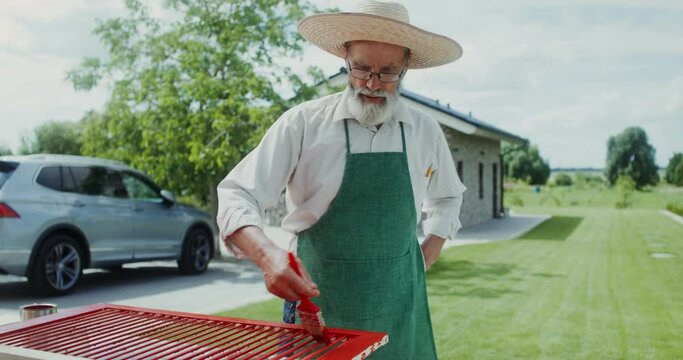 An elderly man in an apron and hat paints the door standing in the backyard of his house